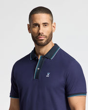 A man with short hair and a beard wears the Psycho Bunny MENS BRANDON PIQUE POLO - B6K280E200, crafted from luxurious cotton pique in navy blue and featuring a logo on the chest. The polo's black collar and light blue trim enhance its elegance as he gazes directly at the camera against a light gray backdrop.