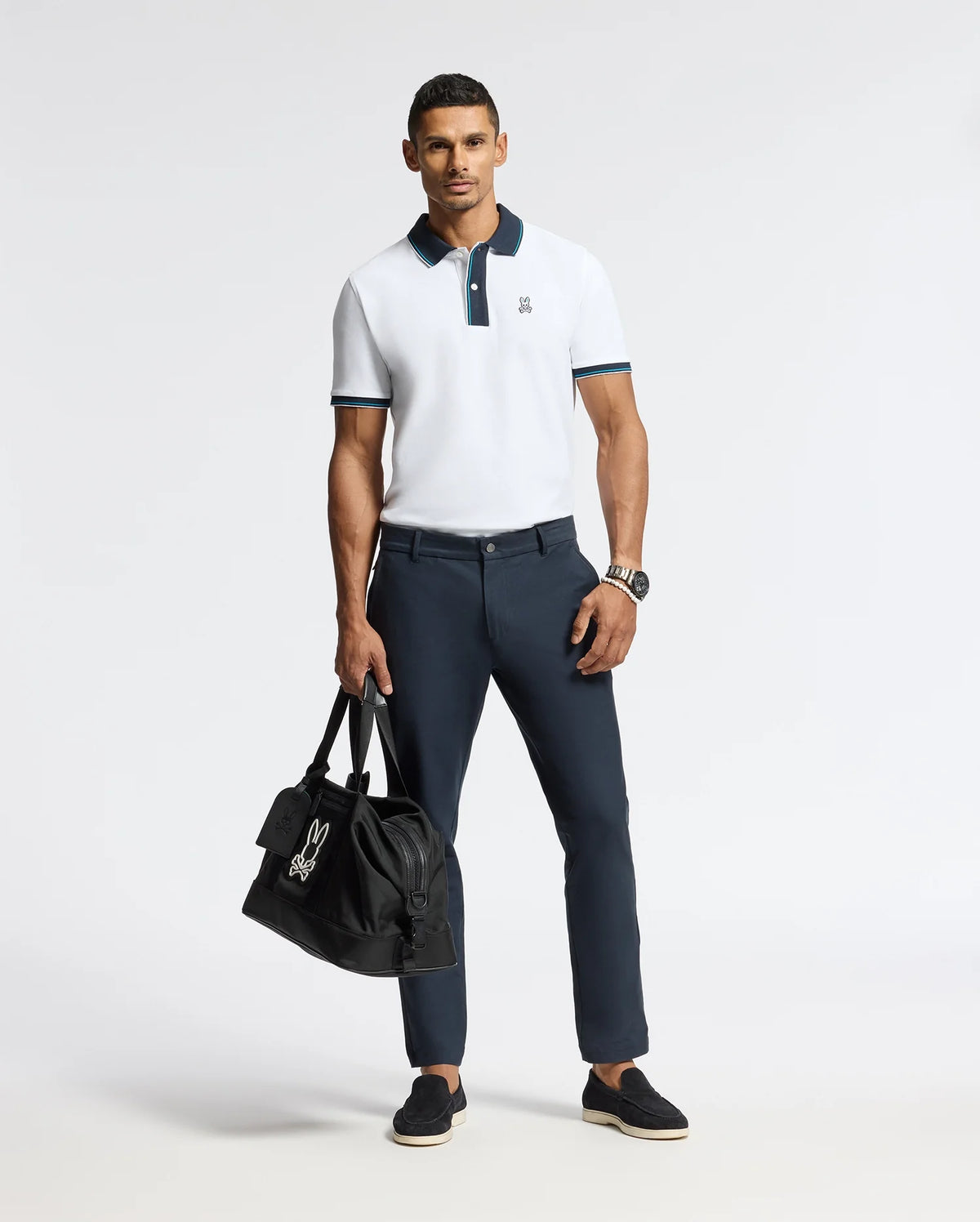 A man poses in a premium quality MENS BRANDON PIQUE POLO (SKU: B6K280E200) from Psycho Bunny and dark trousers, holding a black bag. He wears black shoes and a watch, standing against a plain white background.