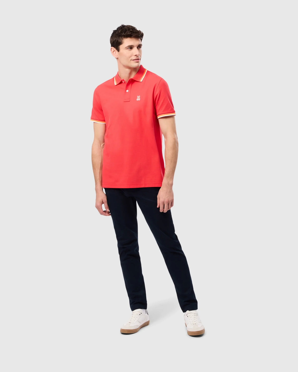 A man is standing against a plain background wearing the Psycho Bunny MENS GRANBURY PIQUE POLO (B6K400C200), which features a tipping collar in red and yellow trim, paired with dark blue pants and white sneakers. His left arm is relaxed by his side while his right arm is slightly bent at the elbow. He is looking to his right.