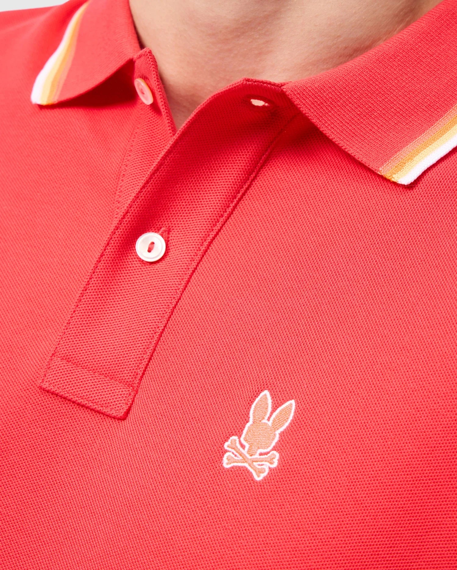 A close-up image of a person wearing the MENS GRANBURY PIQUE POLO - B6K400C200 by Psycho Bunny, showcasing its red Pima cotton fabric with white and yellow striped rib knit trim on the collar. The polo shirt features a small embroidered logo of a rabbit head with crossbones just below the buttons.