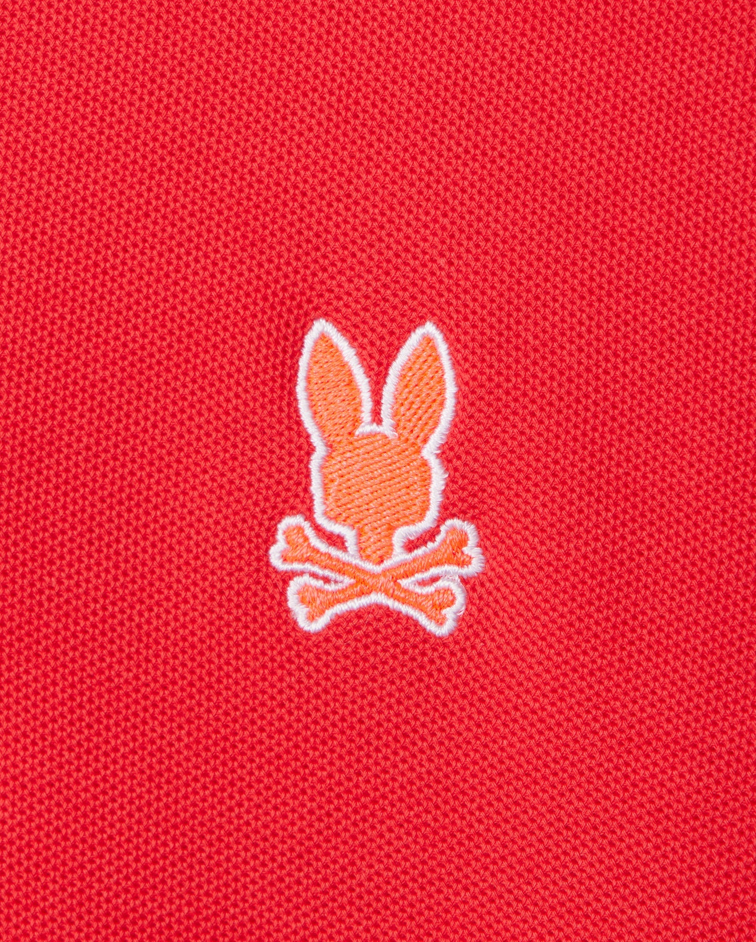 An embroidered orange silhouette of a bunny head with two ears, positioned above a pair of crossed bones in a skull and crossbones motif, adorns the front of the red Pima cotton "MENS GRANBURY PIQUE POLO - B6K400C200" by Psycho Bunny.