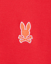 An embroidered orange silhouette of a bunny head with two ears, positioned above a pair of crossed bones in a skull and crossbones motif, adorns the front of the red Pima cotton "MENS GRANBURY PIQUE POLO - B6K400C200" by Psycho Bunny.
