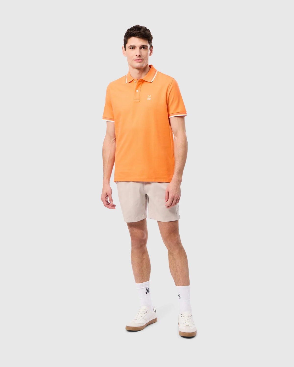 A man wearing an orange Psycho Bunny MENS GRANBURY PIQUE POLO - B6K400C200 crafted from diamond-knit Pima cotton, beige shorts, white socks, and white sneakers stands on a plain light grey background. He is standing in a relaxed pose with his arms by his sides and looking forward.