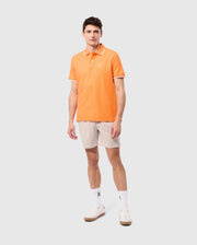 A man wearing an orange Psycho Bunny MENS GRANBURY PIQUE POLO - B6K400C200 crafted from diamond-knit Pima cotton, beige shorts, white socks, and white sneakers stands on a plain light grey background. He is standing in a relaxed pose with his arms by his sides and looking forward.
