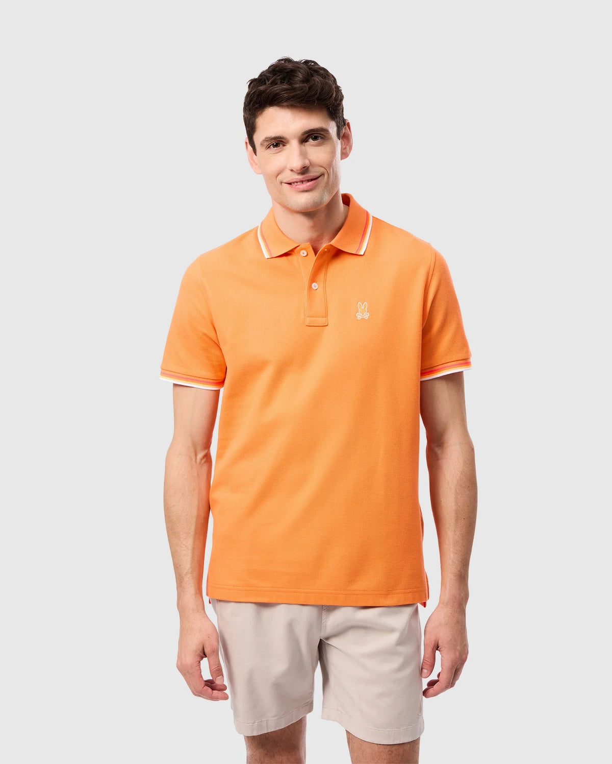 A man with short dark hair is wearing an orange Psycho Bunny MENS GRANBURY PIQUE POLO - B6K400C200 shirt with a diamond-knit Pima cotton design and white trim on the tipping collar and sleeves. The shirt features a small embroidered logo on the left chest. He is also wearing light-colored shorts. The background is plain and light gray.