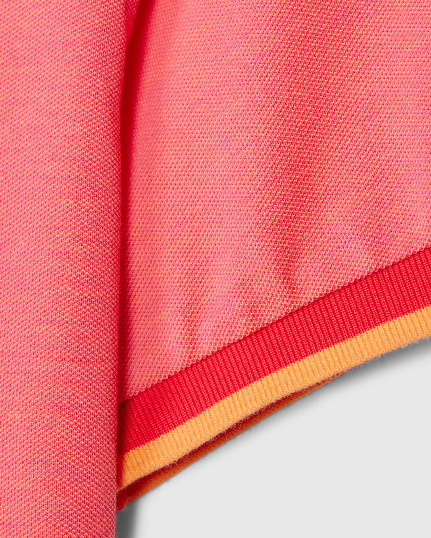 Close-up of the MENS WOODWAY OXFORD POLO by Psycho Bunny, showcasing a pink knit fabric with an elastic hem. The hem features a red band with a thinner orange stripe at the very bottom, crafted from a luxurious Pima cotton-blend. The fabric texture and stitching details are clearly visible.