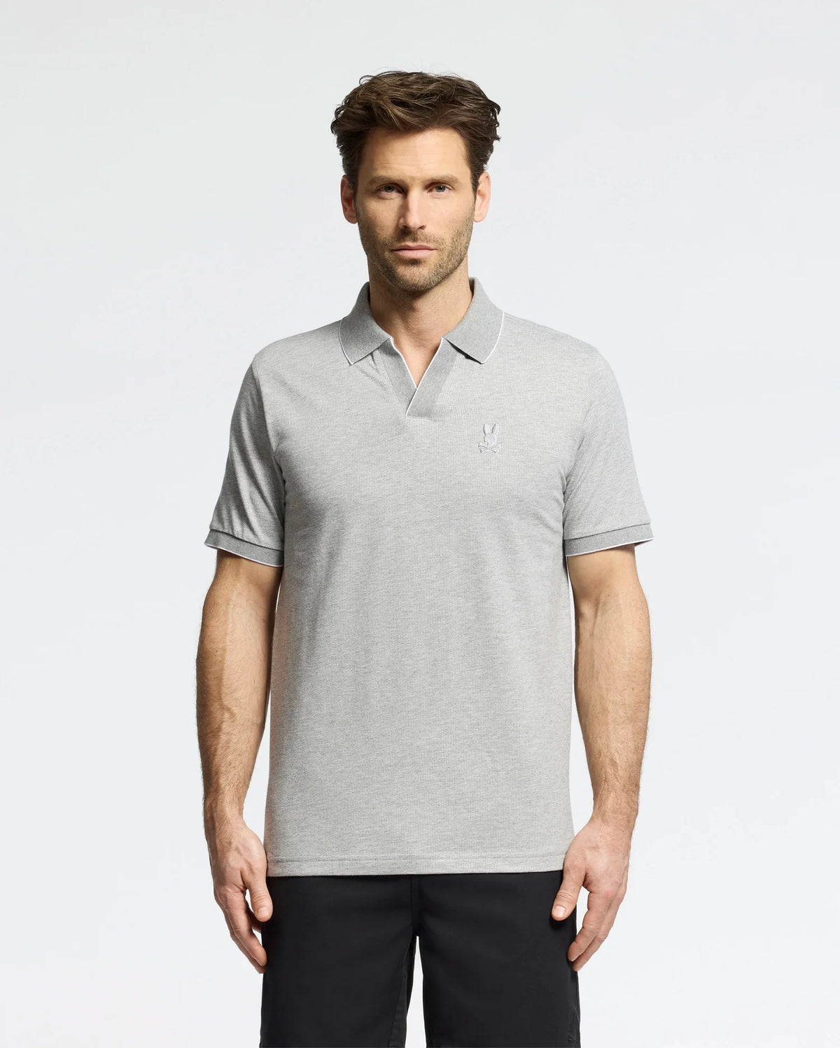 A man in a Psycho Bunny MENS MACKAY JACQUARD JOHNNY COLLAR POLO - B6K408F200, crafted from Pima cotton in light gray with a chest logo, stands against a white background. The polo pairs well with his dark pants. He has short brown hair and a neutral expression.