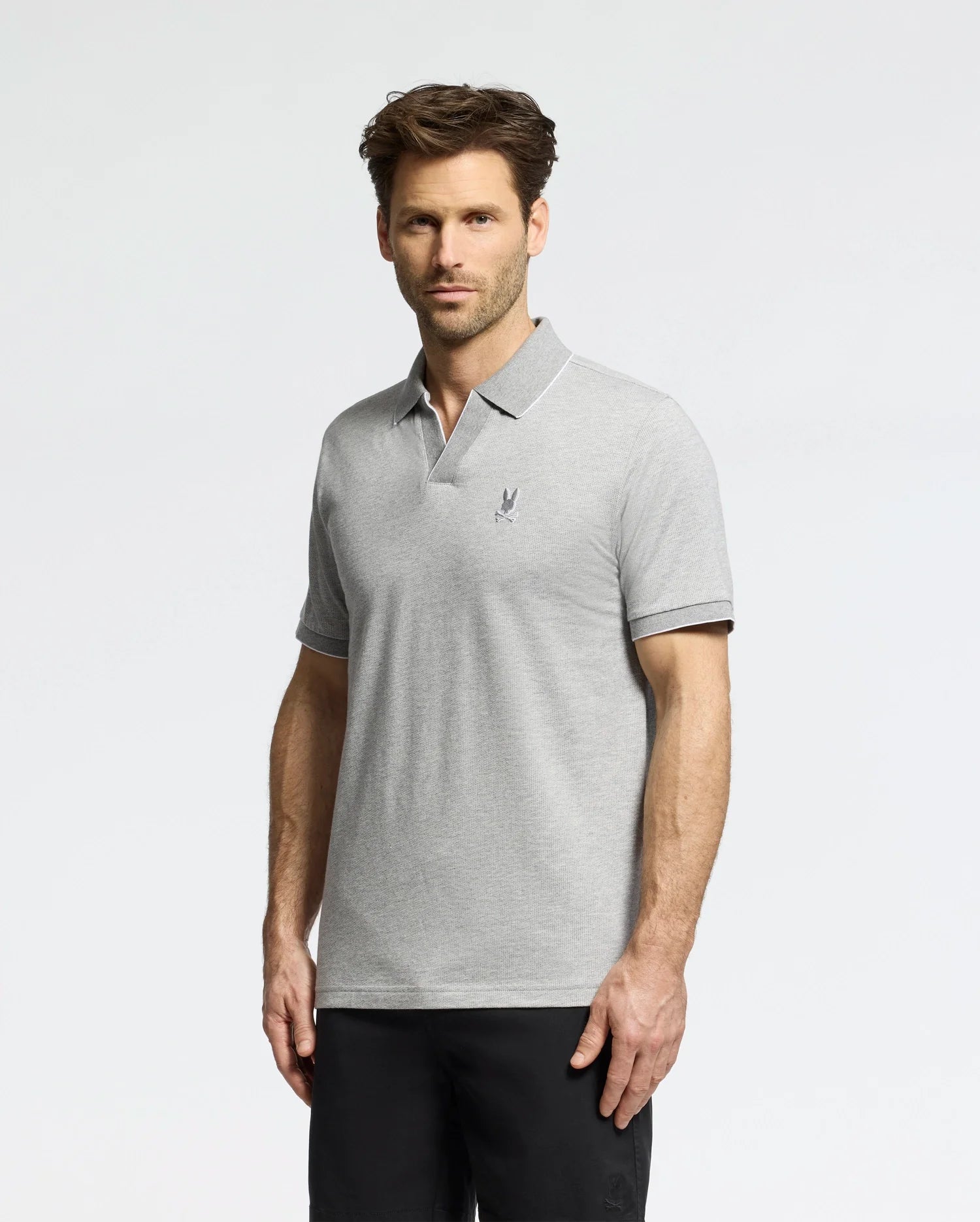 A man with short hair wears the Psycho Bunny MENS MACKAY JACQUARD JOHNNY COLLAR POLO in light gray, featuring a small left chest emblem and classic collar, paired with black pants. He stands against a plain white background, looking directly at the camera with a neutral expression.