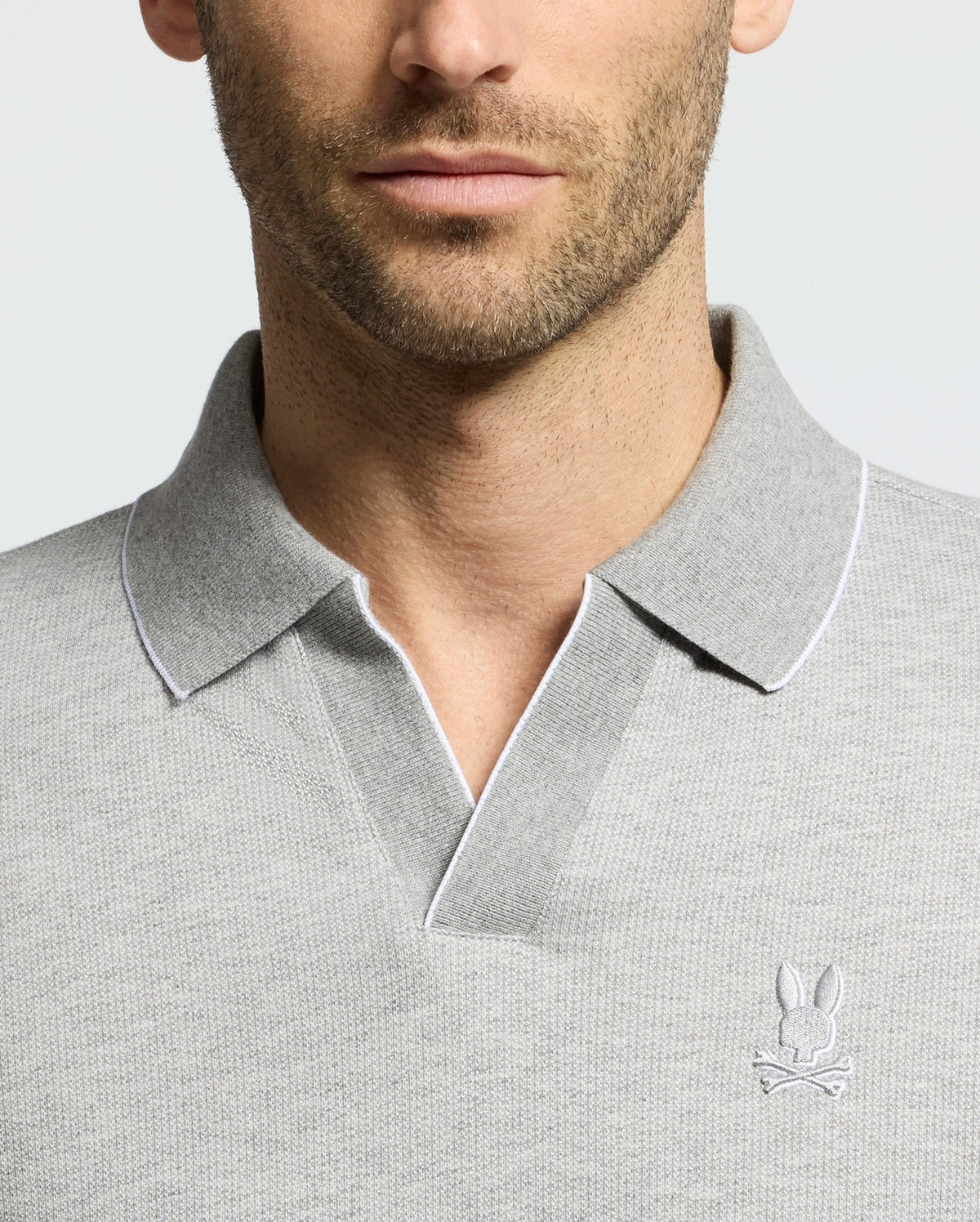 A man wears a Psycho Bunny MENS MACKAY JACQUARD JOHNNY COLLAR POLO with a light gray tone and soft Pima cotton fabric. It features an embroidered bunny and bones logo on the chest, highlighted by white trim. The photo shows his upper torso and part of his face against a neutral backdrop.