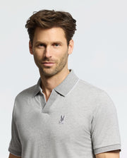 A man in a Psycho Bunny light gray MENS MACKAY JACQUARD JOHNNY COLLAR POLO, made from soft Pima cotton and featuring an embroidered logo on the chest, stands against a plain background, his short brown hair and neutral expression completing the look.