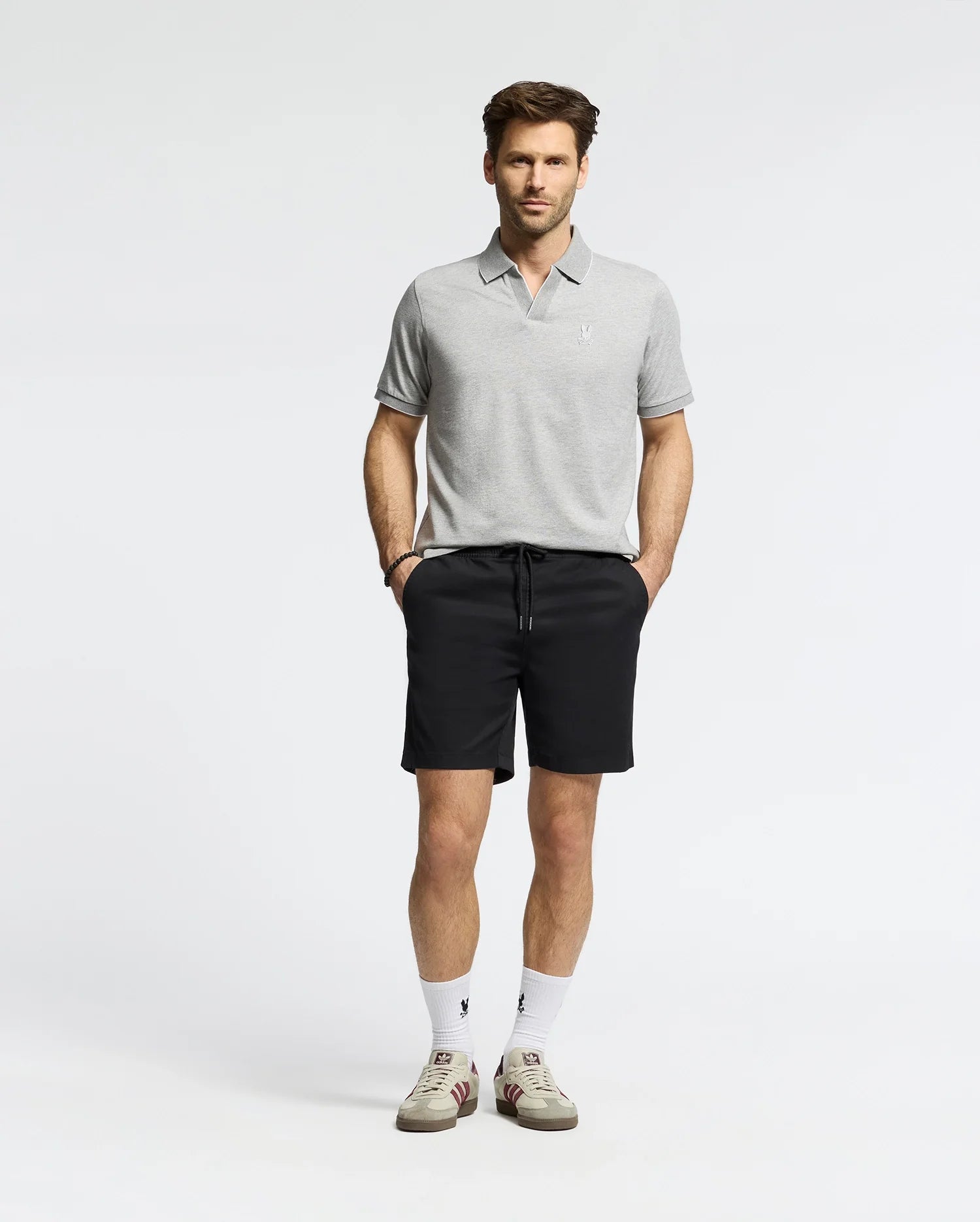 A man wears a Psycho Bunny MENS MACKAY JACQUARD JOHNNY COLLAR POLO (B6K408F200) in gray with black shorts, white socks, and pima cotton sneakers featuring a black "V" logo. He stands against a plain background with hands in pockets and a neutral expression.