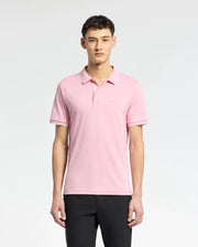 A person stands against a white backdrop wearing Psycho Bunny's Men's Bryce Performance Pique Polo in light pink, made from performance pique fabric with a small chest logo. They have short dark hair and wear black quick-dry, moisture-wicking pants.