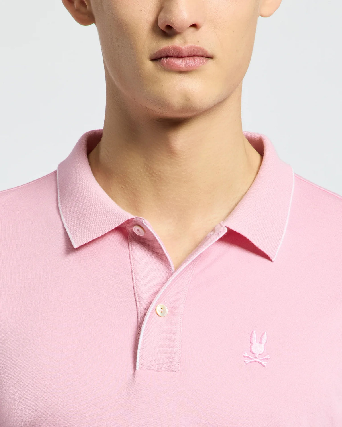 A person wearing a Psycho Bunny MENS BRYCE PERFORMANCE PIQUE POLO - B6K486F200 in light pink, featuring a small bunny logo on the chest. Made from performance pique fabric with quick-dry and moisture-wicking properties, it has a collar and two buttons. The background is plain and neutral.