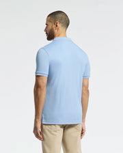 A man with short hair faces away, wearing a Psycho Bunny MENS BRYCE PERFORMANCE PIQUE POLO in light blue and beige pants, set against a plain white background.
