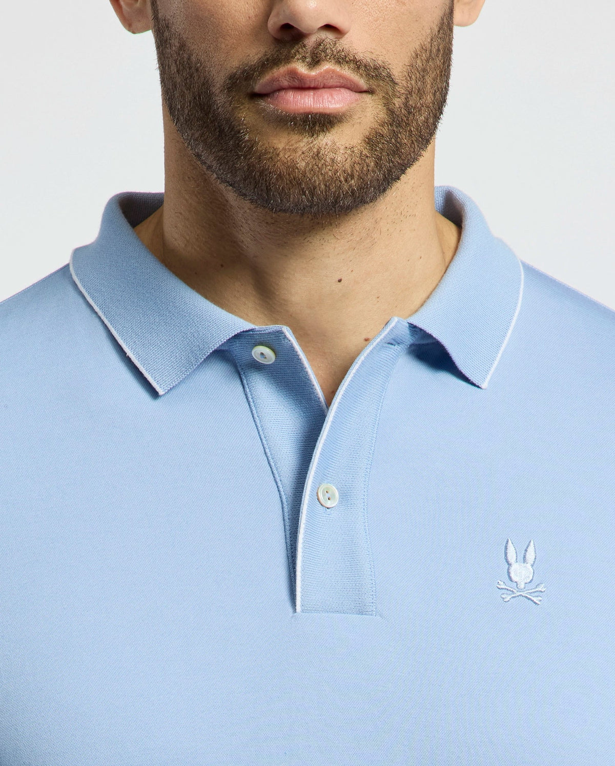 A person wears the Psycho Bunny MENS BRYCE PERFORMANCE PIQUE POLO - B6K486F200 in light blue, featuring a white rabbit and crossed swords emblem. Only the lower face and shoulders are visible, highlighting the shirt's moisture-wicking technology against a plain backdrop.