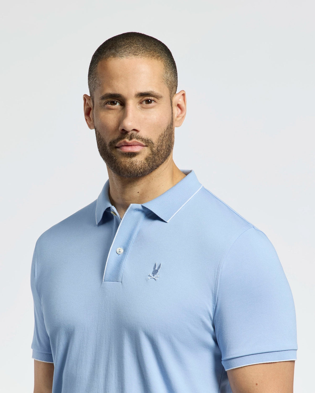 A man with a neatly trimmed beard stands against a plain white background, wearing the Psycho Bunny MENS BRYCE PERFORMANCE PIQUE POLO - B6K486F200 in light blue, which is crafted from moisture-wicking fabric.