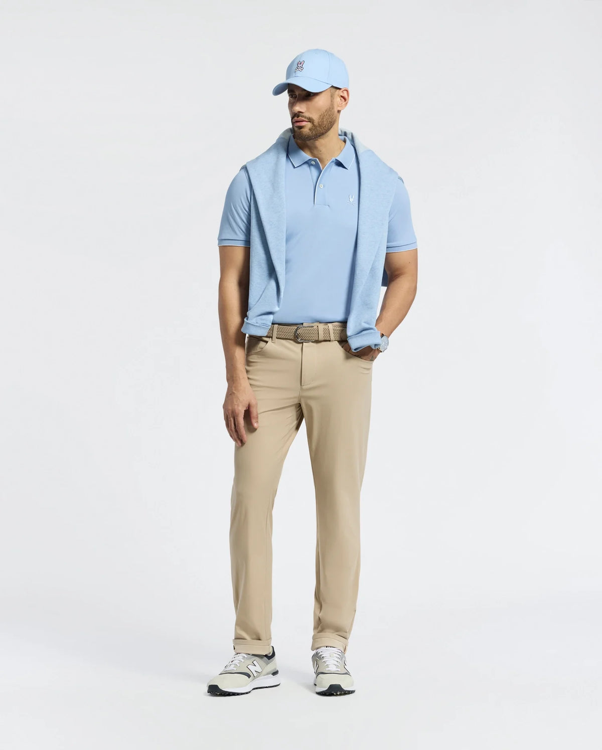 A man stands against a plain white background wearing a Psycho Bunny MENS BRYCE PERFORMANCE PIQUE POLO in light blue, with a matching cap and beige pants. He has a sweater over his shoulders, sports sneakers, wears a belt, and has a watch on his left wrist. The quick-dry fabric ensures comfort.