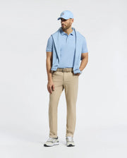 A man stands against a plain white background wearing a Psycho Bunny MENS BRYCE PERFORMANCE PIQUE POLO in light blue, with a matching cap and beige pants. He has a sweater over his shoulders, sports sneakers, wears a belt, and has a watch on his left wrist. The quick-dry fabric ensures comfort.
