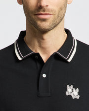 A man dons the Psycho Bunny Men's Elliot Pique Polo (B6K505F200), crafted from soft Pima cotton. The black shirt features a white-striped collar and an embroidered Bunny logo of two bunnies with crossed bones near the collar, set against a plain, light-colored background.