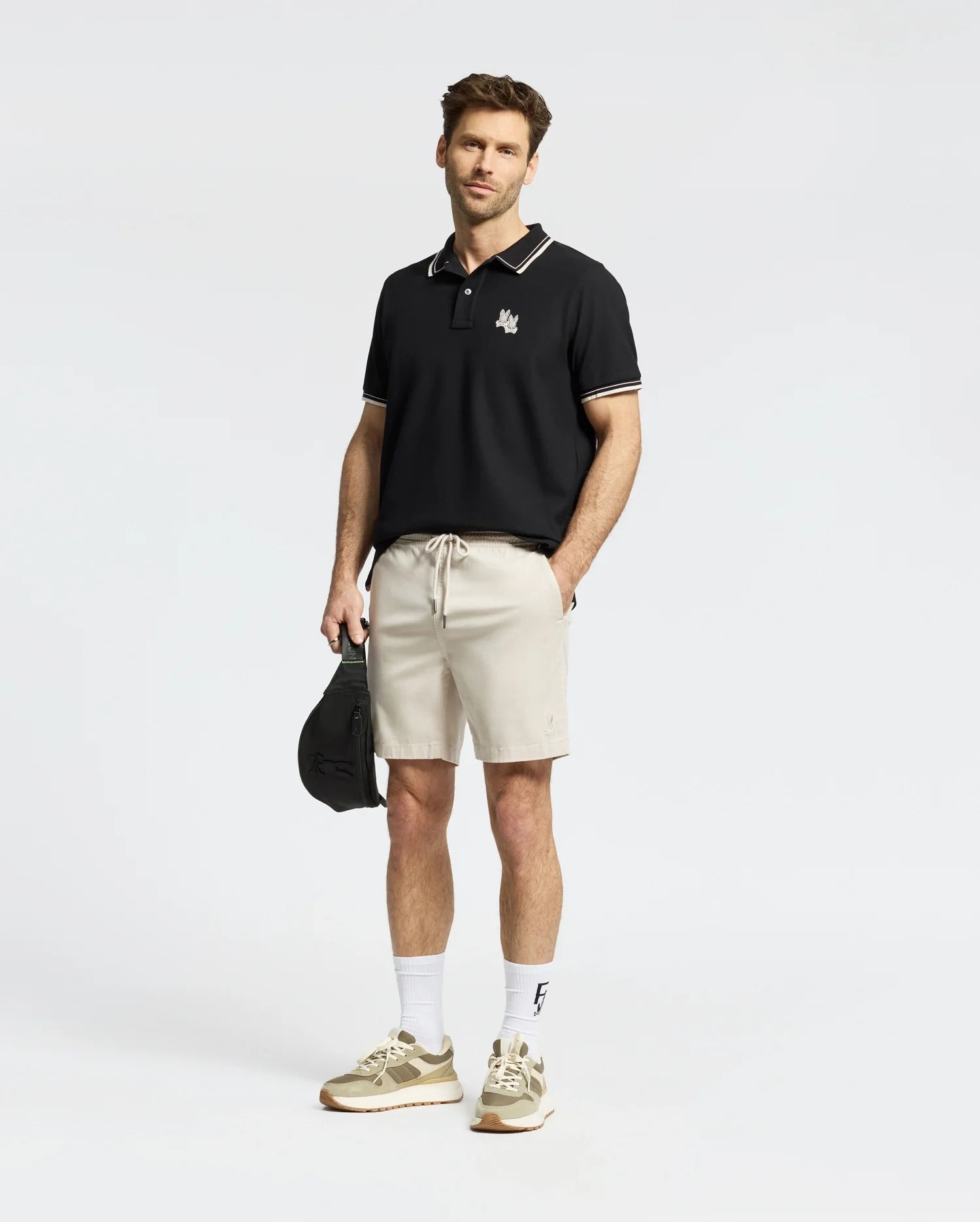 A man with short hair stands against a plain background, wearing a Psycho Bunny men's Elliot Pique polo made of soft Pima cotton and beige shorts. He holds a black cap in his left hand, sporting white socks and beige sneakers.