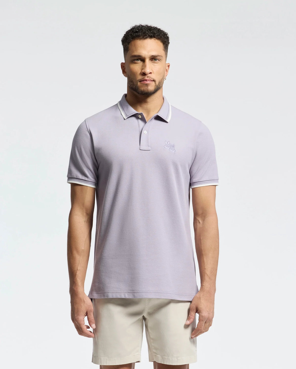 A man with a short beard wears a Psycho Bunny light gray MENS ELLIOT PIQUE POLO - B6K505F200 made of soft Pima cotton with a small chest embroidery, paired with beige shorts. He stands against a plain white background.