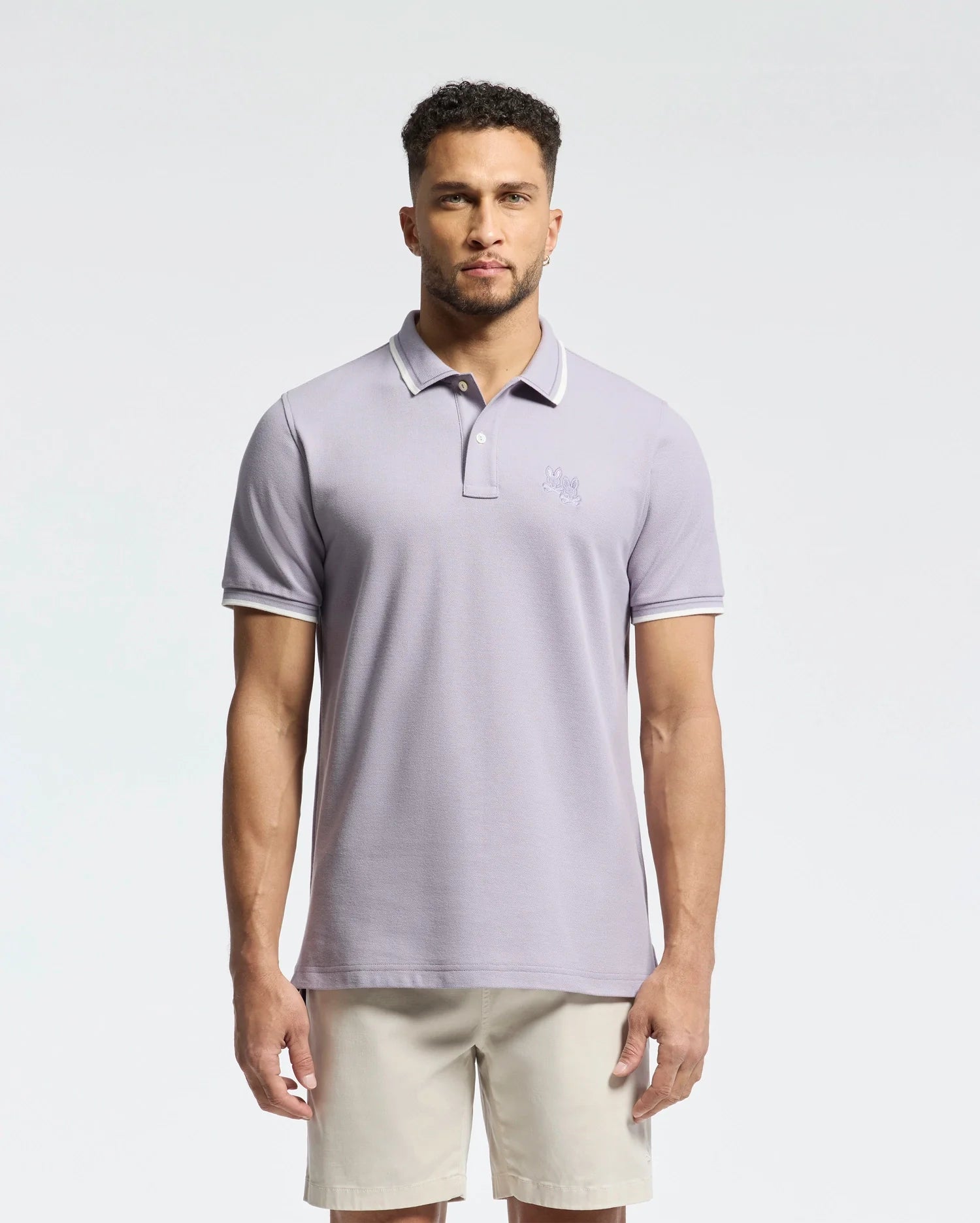 A man with a short beard wears a Psycho Bunny light gray MENS ELLIOT PIQUE POLO - B6K505F200 made of soft Pima cotton with a small chest embroidery, paired with beige shorts. He stands against a plain white background.