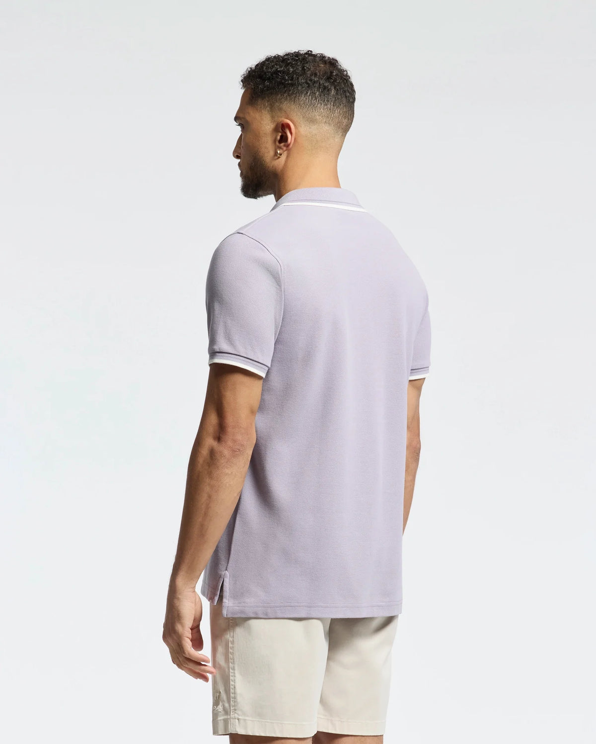 Man with short hair in a light purple Psycho Bunny Men's Elliot Pique Polo (B6K505F200), made from soft Pima cotton, paired with beige shorts, standing and facing left against a plain white background.