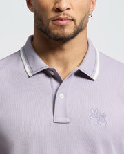 The Psycho Bunny MENS ELLIOT PIQUE POLO (B6K505F200) is crafted from Pima cotton in a light purple with a white-trim collar, featuring two cartoonish rabbit heads embroidered on the left chest for a bold design against a neutral backdrop.