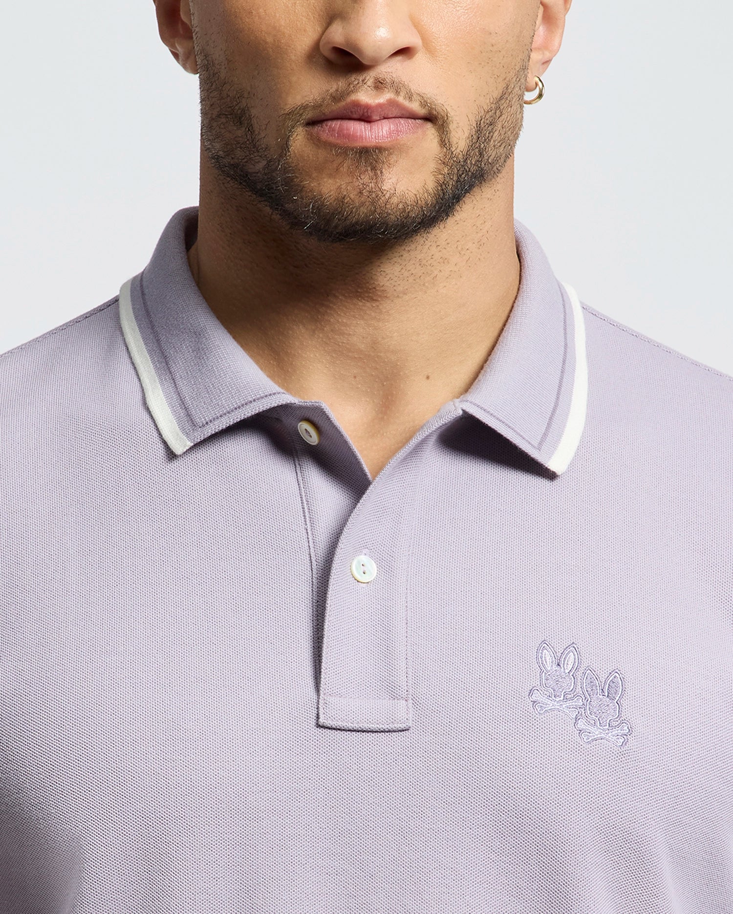 The Psycho Bunny MENS ELLIOT PIQUE POLO (B6K505F200) is crafted from Pima cotton in a light purple with a white-trim collar, featuring two cartoonish rabbit heads embroidered on the left chest for a bold design against a neutral backdrop.