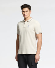 A man in a Psycho Bunny MENS ELLIOT PIQUE POLO - B6K505F200, a light beige shade with soft Pima cotton and subtle chest logo, paired with dark pants against a plain white background.