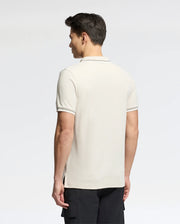 Facing away, a man dons a Psycho Bunny Men's Elliot Pique Polo (B6K505F200) made from soft Pima cotton. The shirt is light-colored with bold dark trim on the sleeves and collar, set against a plain, light gray background.