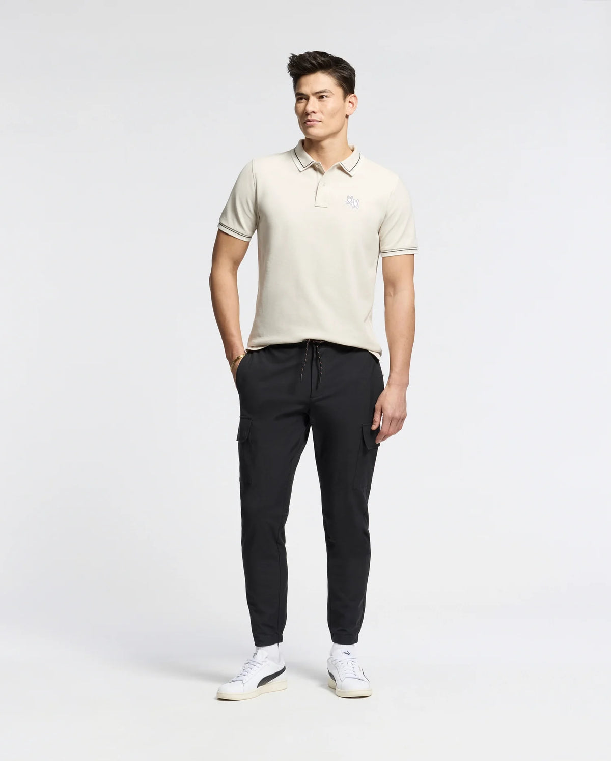 Against a plain backdrop, a man wears the Psycho Bunny MENS ELLIOT PIQUE POLO - B6K505F200 crafted from Pima cotton. He complements it with dark jogger pants and white sneakers, hands in pockets, looking sideways with effortless style.