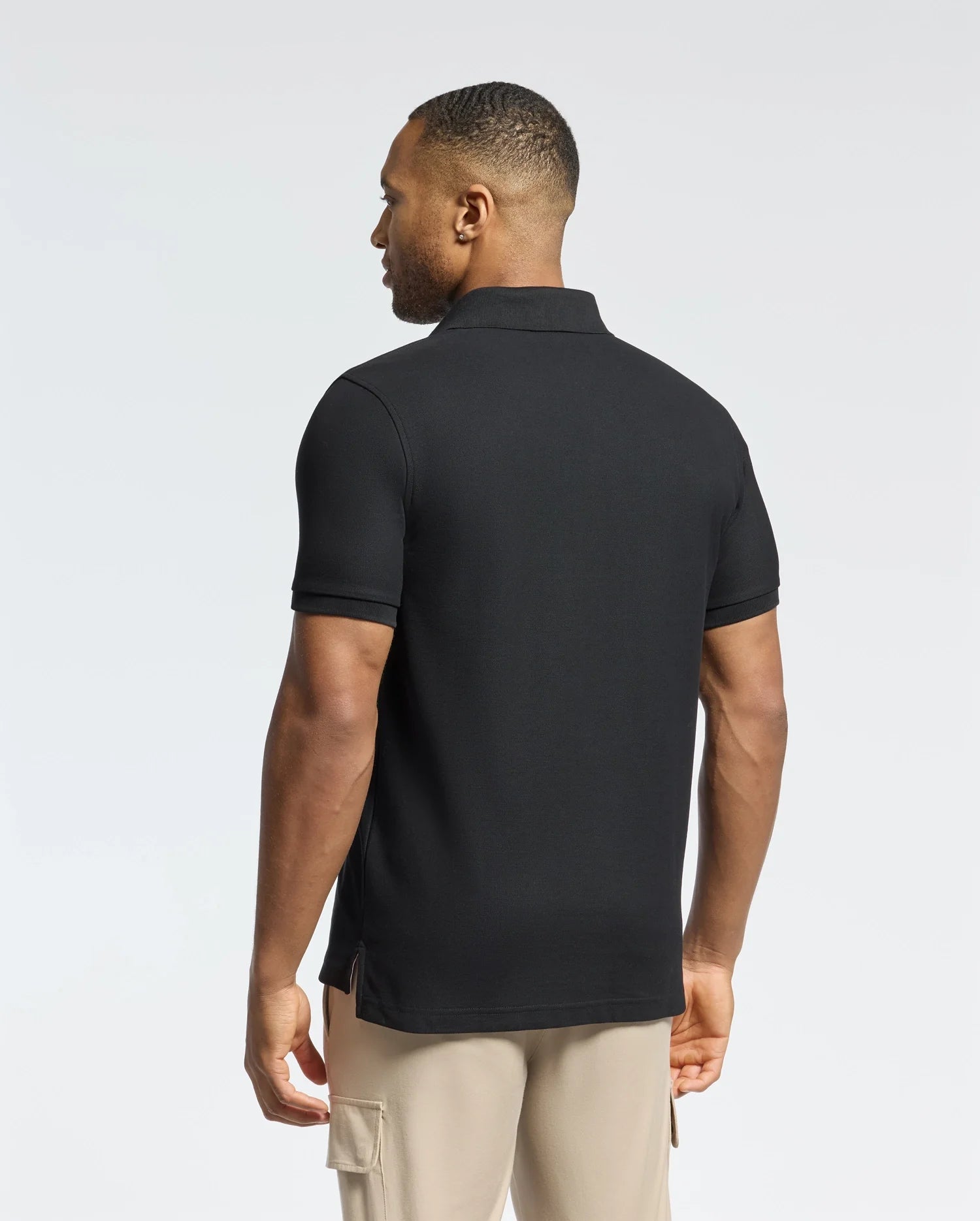 A man wearing the Psycho Bunny MENS DESERT PIQUE POLO - B6K506F200 made from premium Pima cotton along with beige pants is facing away, looking to his left. The plain white background highlights his timeless style.
