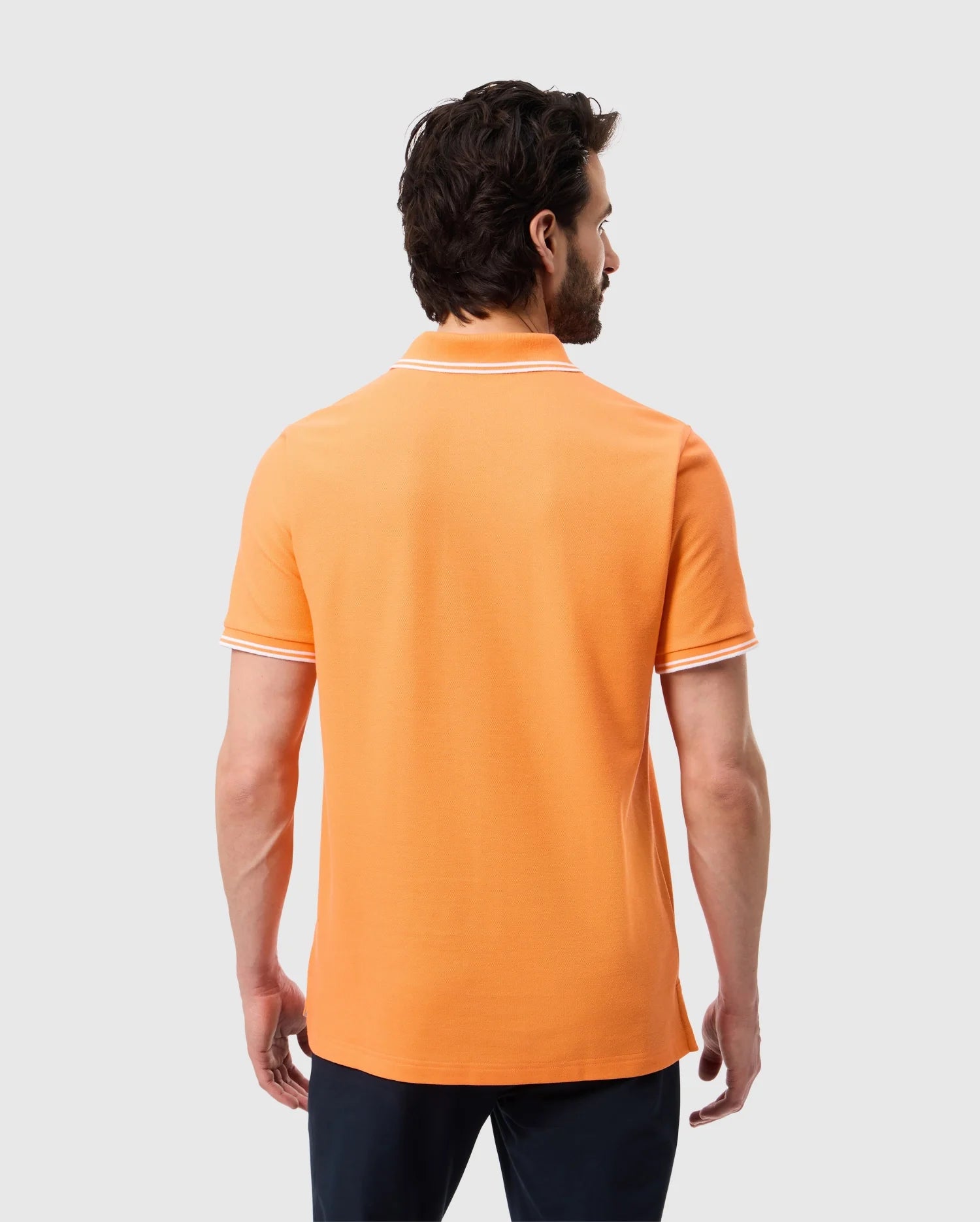 A man with dark hair and a beard is standing with his back to the camera. He is wearing an orange Psycho Bunny MENS BELTON PIQUE POLO - B6K575C200 made of soft Pima cotton, featuring white trim on the collar and sleeves, paired with dark pants. The background is plain white.