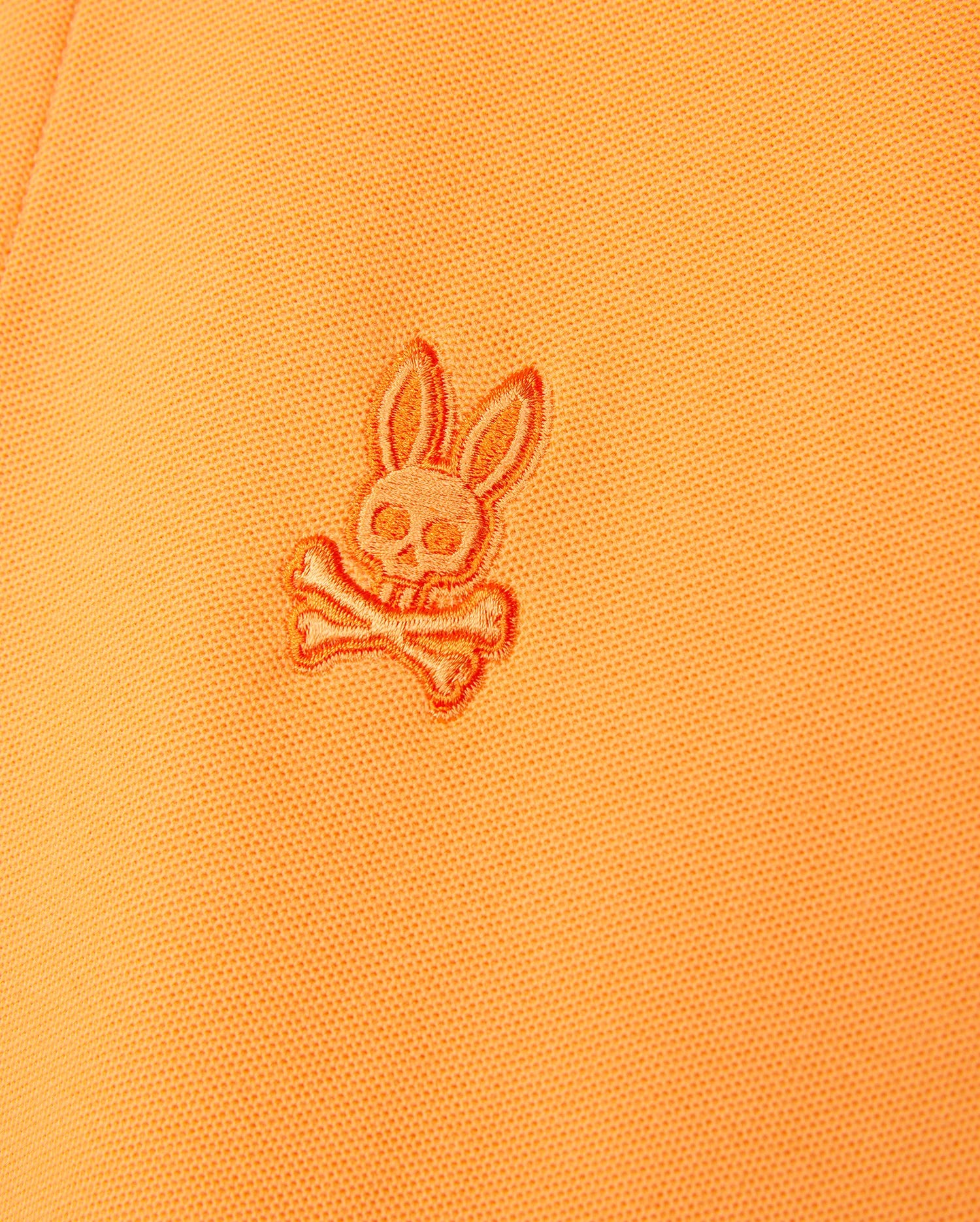 Close-up of an orange Psycho Bunny MENS BELTON PIQUE POLO - B6K575C200 made from Pima cotton featuring a stitched embroidered Bunny logo. The detailed embroidery showcases a stylized bunny skull with large ears above two crossed bones.