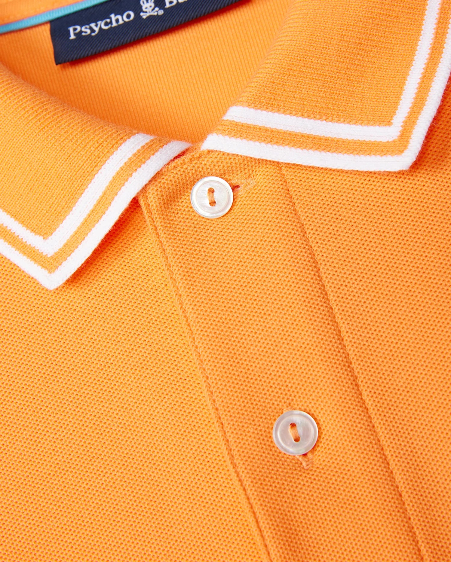 Close-up of a MENS BELTON PIQUE POLO - B6K575C200 showing part of the collar and buttons. The collar has white piping along the edges. A label from the brand "Psycho Bunny" is partially visible at the top, accompanied by their embroidered Bunny logo. The shirt fabric, made from Pima cotton, has a textured appearance.