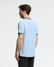A man, with short brown hair, models a Psycho Bunny MENS JONAS MICRO STRIPE COLLAR PIQUE POLO in light blue with dark collar trim, made of premium Pima cotton. He pairs it with black pants against a plain, light-colored background.