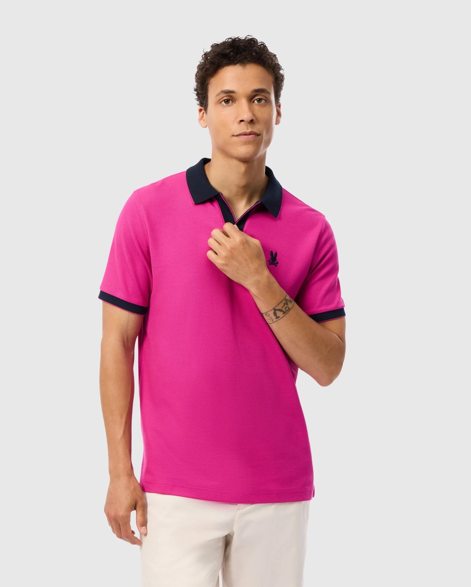 A young man with short, curly hair is wearing a bright pink Psycho Bunny MENS RICHARDS JOHNNY COLLAR PIQUE POLO SHIRT - B6K641C200, featuring black collar and sleeve trim. Paired with white pants, he stands against a plain white background, lightly holding the shirt collar with his right hand. The breathable Pima cotton ensures comfort while displaying a tattoo on his left forearm.
