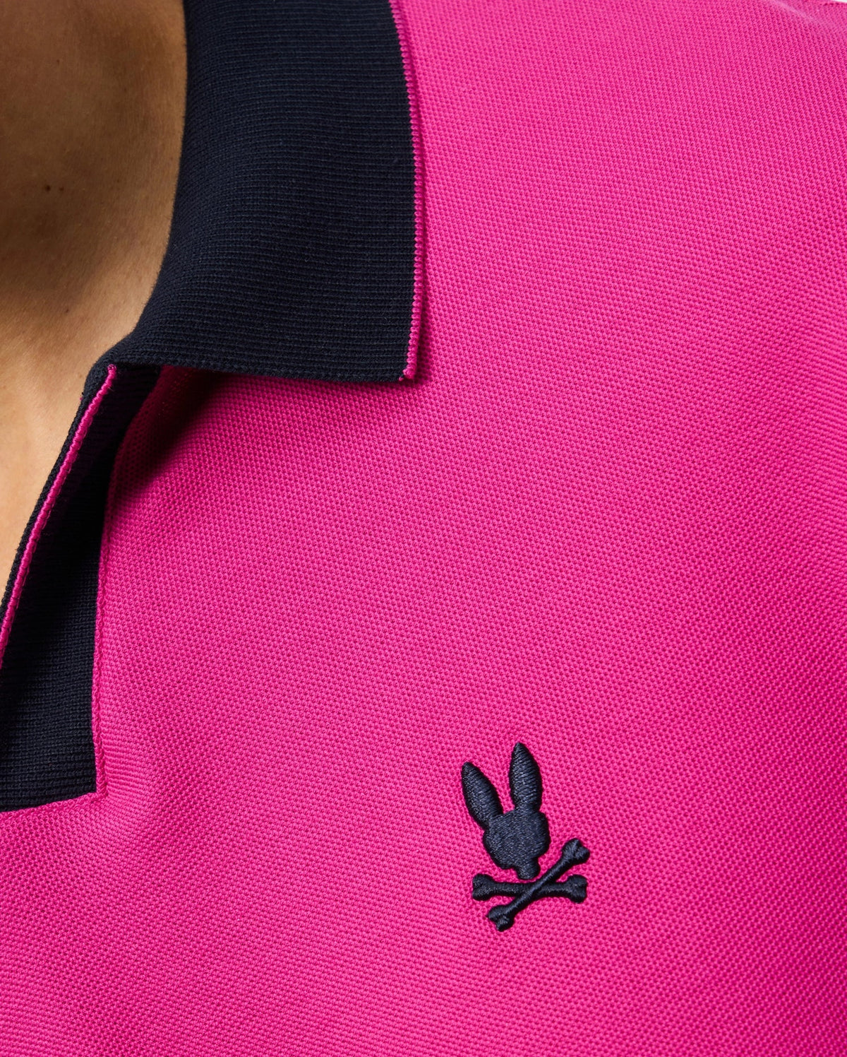 Close-up of a person wearing the Psycho Bunny Men's Richards Johnny Collar Pique Polo Shirt (B6K641C200) in pink, featuring a contrasting black collar. This breathable Pima cotton polo showcases an embroidered black logo on the left side of the chest, depicting a rabbit head with crossed bones. Part of the person's neck and shoulder are visible.