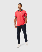 A smiling man wearing a bright red MENS KAYDEN PIQUE POLO - B6K675C200 by Psycho Bunny with navy collar details, navy pants, and white sneakers. He has one hand in his pocket and stands against a plain light gray background.