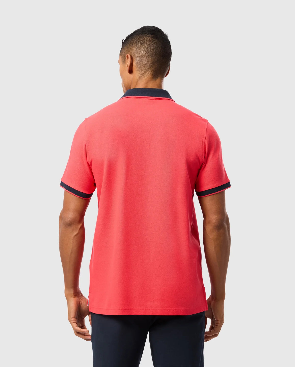 A man is standing with his back to the camera, wearing a bright red Psycho Bunny MENS KAYDEN PIQUE POLO - B6K675C200 with contrasting stripes on the collar and sleeve edges. He has short hair and is also wearing dark navy trousers. The background is plain light gray.