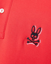 Close-up of a red Psycho Bunny MENS KAYDEN PIQUE POLO - B6K675C200 featuring a small embroidered logo on the left side. The logo consists of a black skull with rabbit ears above a pair of crossbones. The shirt, made from diamond-knit Pima cotton, also has a partially visible button and collar.