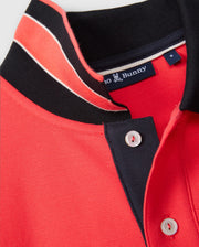 Close-up of a red MENS KAYDEN PIQUE POLO - B6K675C200 with black and white contrasting stripes on the collar and button placket. The brand label inside the collar reads "Psycho Bunny" and features a small bunny logo. Made from diamond-knit Pima cotton, the shirt is neatly folded at the collar.