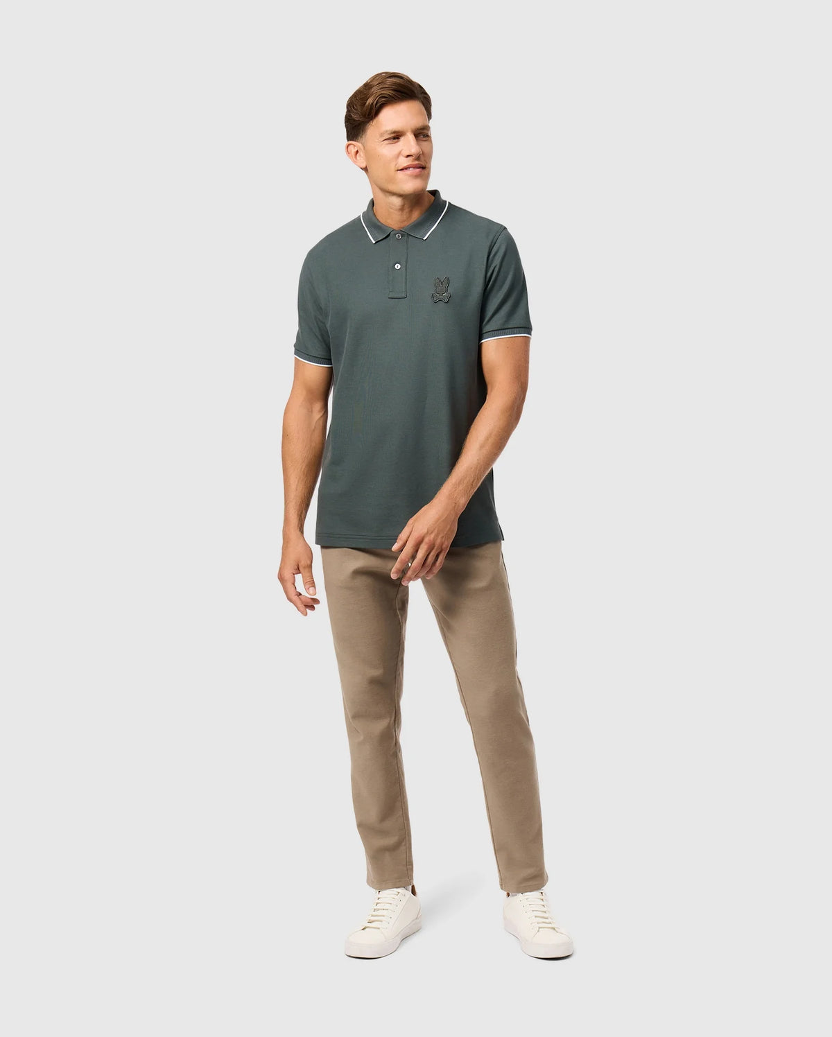 A man stands confidently against a plain background, wearing a dark green Mens Kingston Pique Polo Shirt (B6K764D200) by Psycho Bunny, featuring an embroidered Bunny patch on the chest. He pairs it with beige pants and white sneakers. The Pima cotton fabric of the polo shirt adds a touch of casual elegance. He has short hair and shows a slight smile, with his left hand relaxed at his side and his right hand slightly bent.