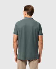 A man with short hair is standing with his back to the camera, wearing a green Psycho Bunny MENS KINGSTON PIQUE POLO SHIRT - B6K764D200 made of Pima cotton and beige pants. The plain, light-colored background highlights his casual attire and the embroidered Bunny patch on his shirt.
