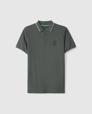 The Psycho Bunny MENS KINGSTON PIQUE POLO SHIRT - B6K764D200 is a dark green polo shirt made from luxurious Pima cotton. It features a collar and short sleeves with white trim along the edges, and an embroidered Bunny patch on the left chest. The shirt is displayed against a plain white background.