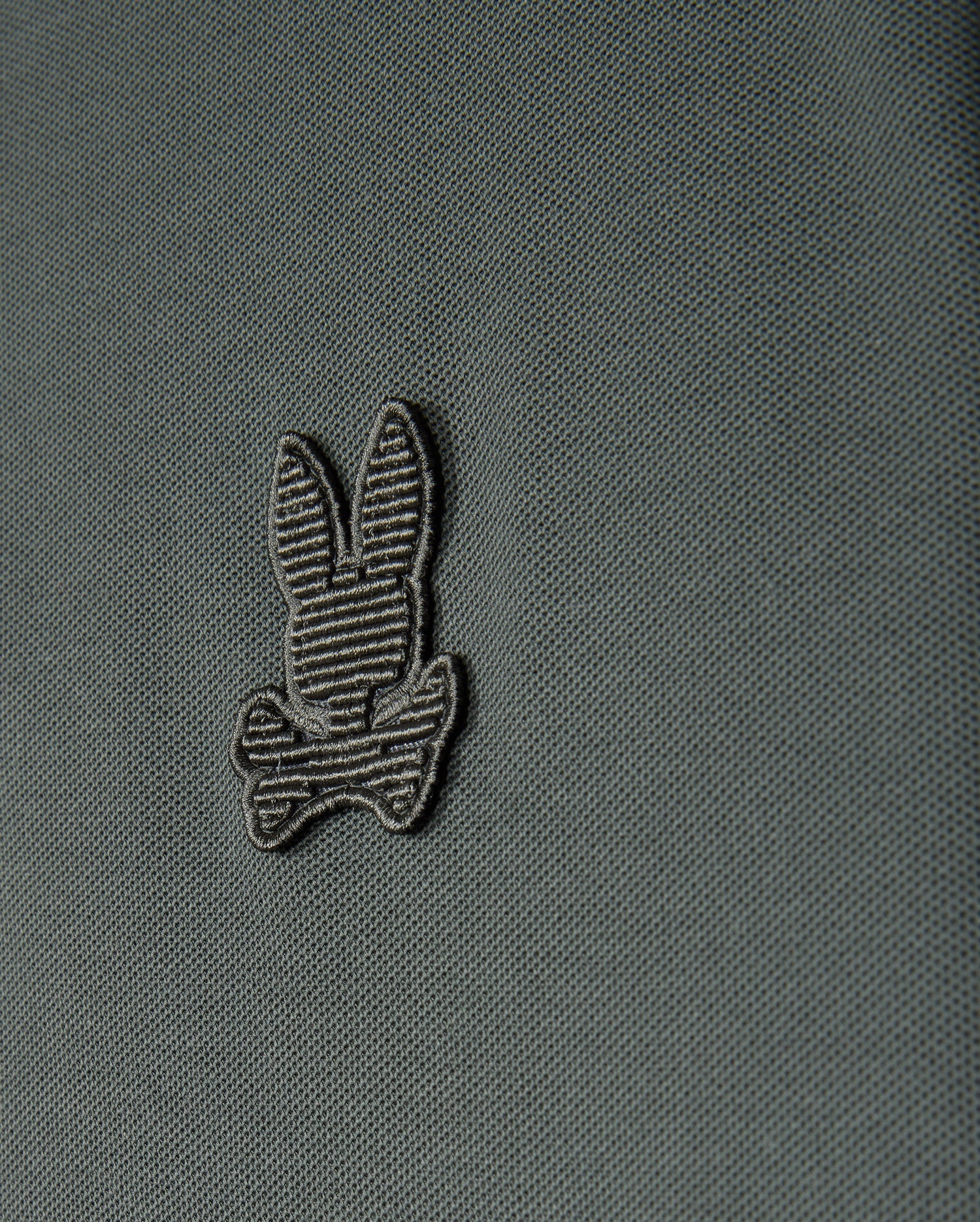 A close-up view of the Psycho Bunny Men's Kingston Pique Polo Shirt (B6K764D200) showcases an embroidered Bunny patch featuring a striped bunny head above two crossed bones, reminiscent of the Jolly Roger flag. The design is stitched with darker thread that subtly contrasts with the fabric.