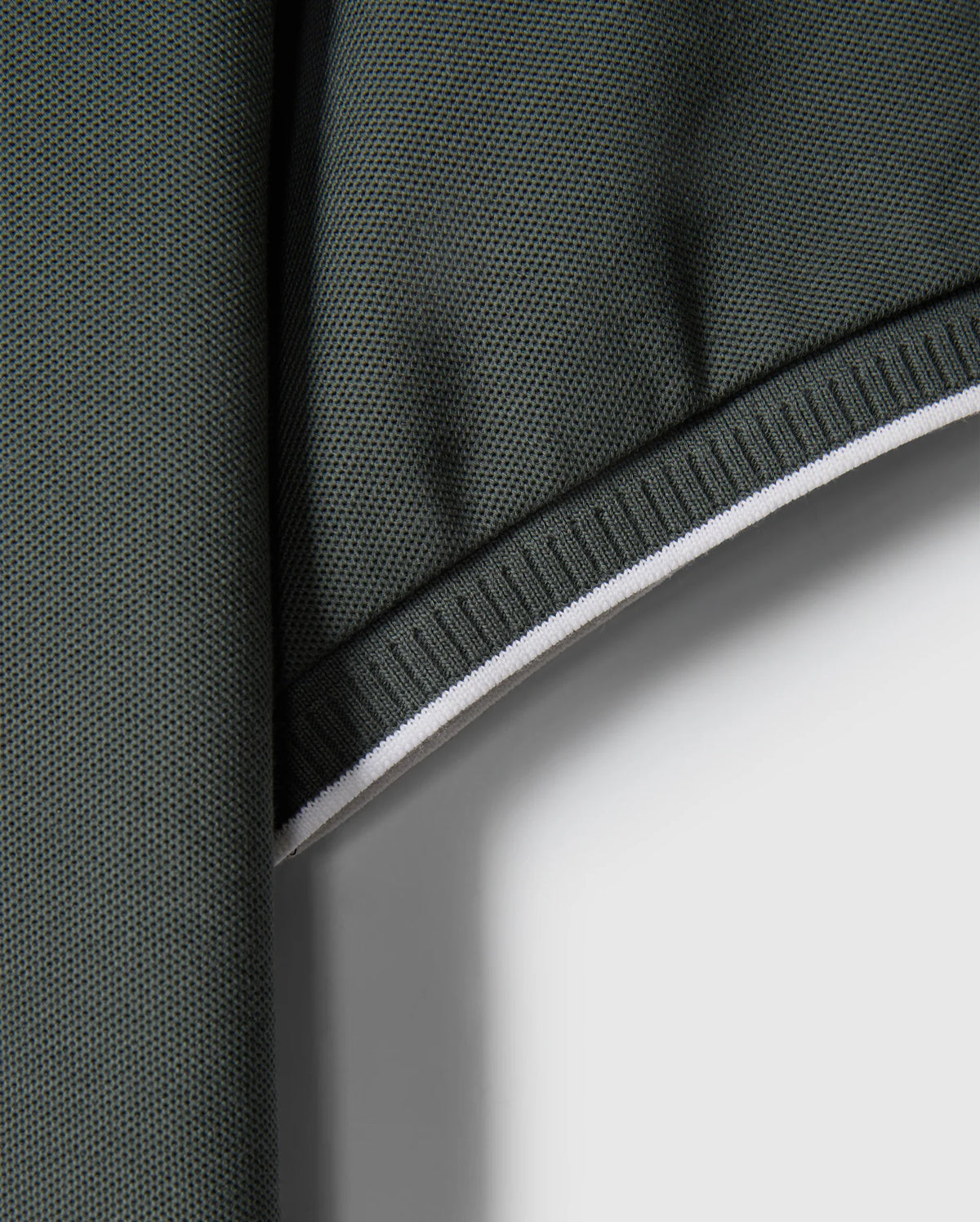 Close-up of a green Pima cotton fabric with a textured surface and white edge. The fabric appears to be folded at the top, creating a shadowed area. The edge is reinforced with white stitching, providing contrast against the dark green material – perfect for crafting a stylish MENS KINGSTON PIQUE POLO SHIRT - B6K764D200 by Psycho Bunny, perhaps with an embroidered Bunny patch.