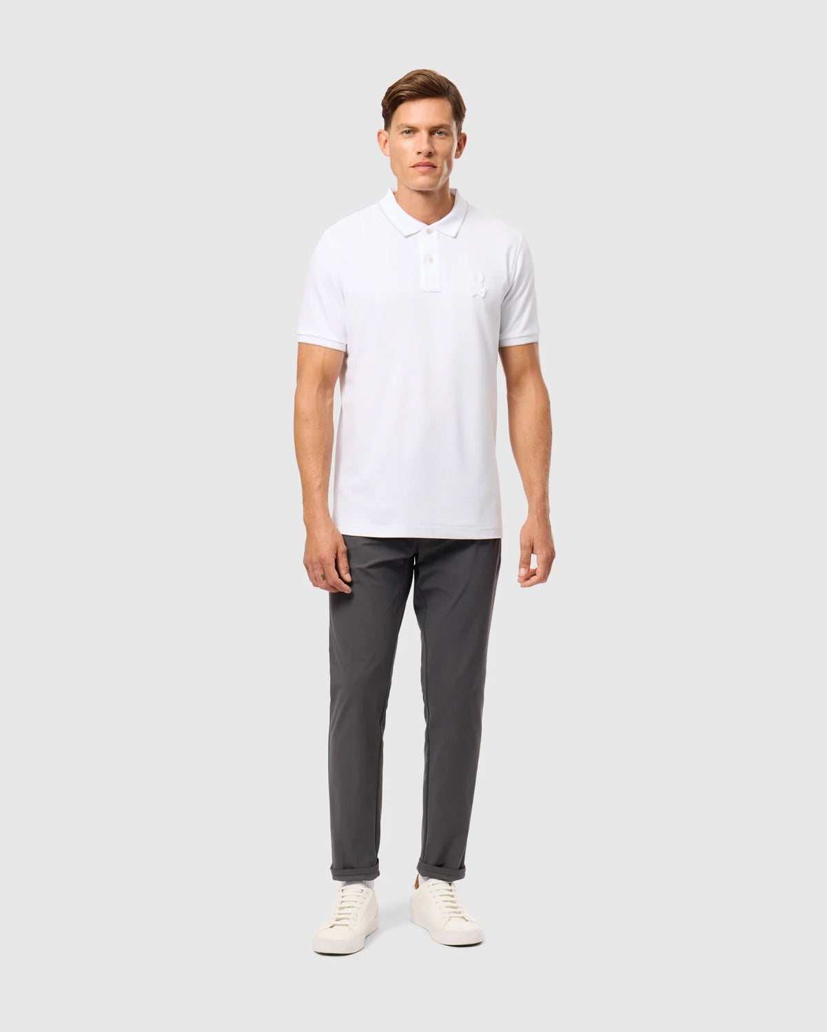 A man stands against a plain background, wearing a white Psycho Bunny Mens Kingston Pique Polo Shirt (B6K764D200) with an embroidered Bunny patch, dark gray pants, and white sneakers. He has short hair and is posing with his arms by his sides, looking straight ahead.