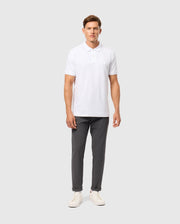 A man stands against a plain background, wearing a white Psycho Bunny Mens Kingston Pique Polo Shirt (B6K764D200) with an embroidered Bunny patch, dark gray pants, and white sneakers. He has short hair and is posing with his arms by his sides, looking straight ahead.
